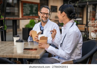 Two doctors wearing white lab coats and stethoscopes are sharing a pleasant outdoor lunch break, eating from take-out boxes at a restaurant table while smiling and talking together