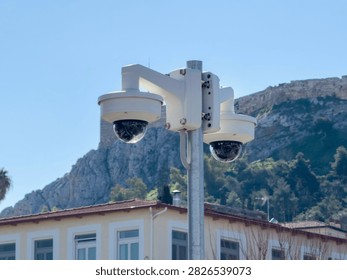 Two dome security cameras on a metal pole monitor buildings below a rocky hillside in Monastiraki, Athens, Greece, blue sky