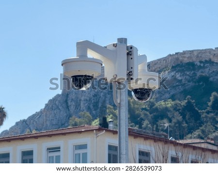 Two dome security cameras on a metal pole monitor buildings below a rocky hillside in Monastiraki, Athens, Greece, blue sky