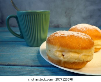 Two Doughnuts, Sprinkled With Powdered Sugar, On A White Plate Next To A Dark Mug Of Coffee On A Blue Wooden Table. Side View