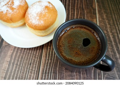 Two Doughnuts, Sprinkled With Powdered Sugar, Lay On A White Plate Next To A Dark Coffee Mug On A Wooden Table. Side View