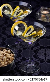 Two dry martini cocktails with a slice of lemon by a bowl of pistachios and a candle over a purple tablecloth.