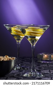 Two dry martini cocktails with a slice of lemon by a bowl of pistachios and a candle over a purple tablecloth, purple night light in the background.