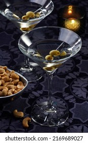 Two dry martinis with olives by a bowl of cashew nuts and a candle over a purple tablecloth.