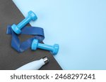 Two dumbbells, yoga mat, fitness elastic band and thermo bottle on light blue background, flat lay. Space for text