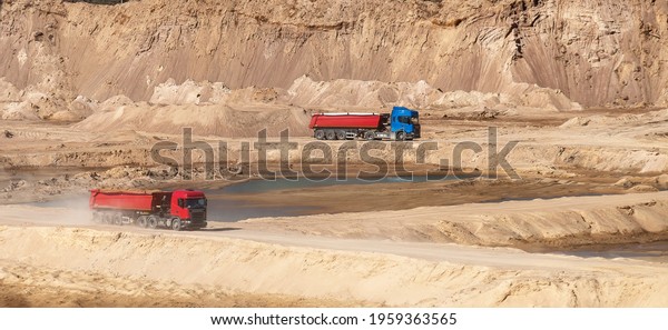 A two dump trucks with semi-trailers ride against\
the background of a sand pit (quarry, dunes), raising dust behind\
it. Wide 16:9 image.