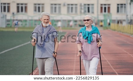 Two elder aged adults practice nordic walk poles. Old senior caucasians couple trekking sticks hike. Healthy love smile lifestyle. Old grandparent hikers couples workout stadium. Elderly sport people.