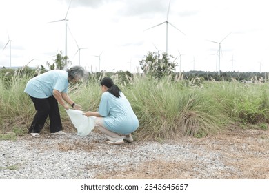 Two elderly Asian woman volunteers collecting trash at wind turbine farm. Volunteer cleans up the meadow with garbage collection with wind turbines park at background. Ecology and environment concept - Powered by Shutterstock