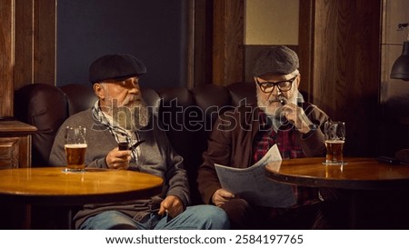 Two elderly bearded men in caps sitting in leather armchairs, smoking tobacco pipes, reading newspaper and drinking bee in cozy bar. Concept of elderly people lifestyle, drinks, retro fashion