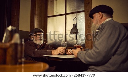 Two elderly bearded men in retro-style clothes with caps, drinking beer at wooden table in rustic atmospheric bar, sharing casual and friendly moment. Concept of elderly people lifestyle, drinks