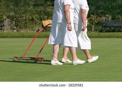 Two Elderly Females In White Bowling Clothes Walking Together On A Green Lawn, One Of The Females Pulling A Red Metal Ball Gatherer.