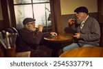 Two elderly men drinking beer at wooden table in rustic atmospheric bar, sharing casual and friendly moment. Concept of elderly people lifestyle, drinks, retro fashion