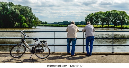 Two elderly men leaning over a bridge railing and looking musing over the water. Next to them an e-bike is parked.