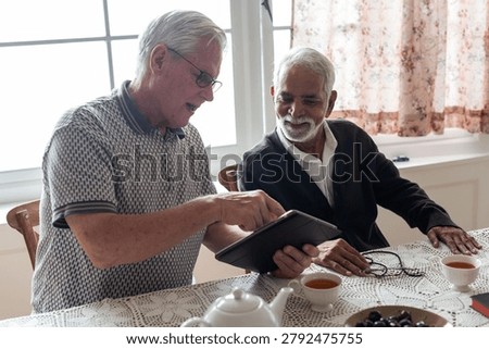 Two elderly men, one white and one South Asian, sit at a dining table sharing a tablet and chatting over tea in a bright room.