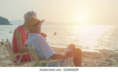 Two elderly men and women sit chair at the beach talking and watching the sun and the sea on their summer vacation and they smile and enjoy their vacation. - Powered by Shutterstock