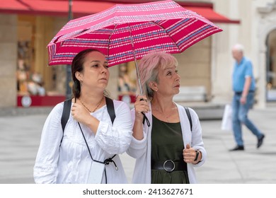 Two elderly and middle-aged women are walking under an umbrella in a European city. Concept: tourism and travel, female friendship, family relations. - Powered by Shutterstock