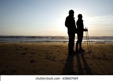 Two Elderly People Standing On A Beach At Egmond, The Netherlands. One Person Is Nordic Walking. They Are Silhouettes Against The Background Of The Setting Sun.