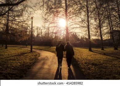 Two Elderly People Walking In A Park In The Sunset