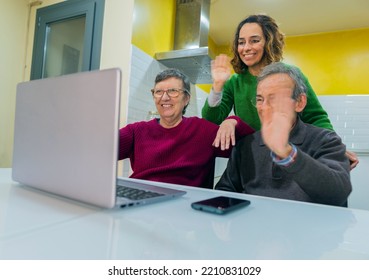 Two Elderly People And A Woman Waving Through At A Laptop Screen In The Kitchen Of Their Home