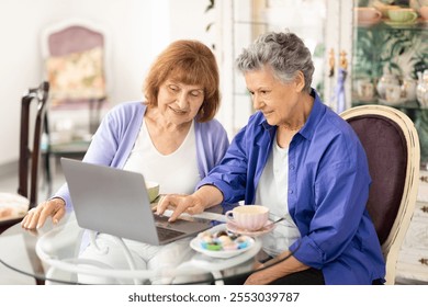 Two elderly women share a relaxing moment at a cafe, engaged in lively conversation while browsing on a laptop. They enjoy tea and colorful pastries, embodying a joyful lifestyle. - Powered by Shutterstock