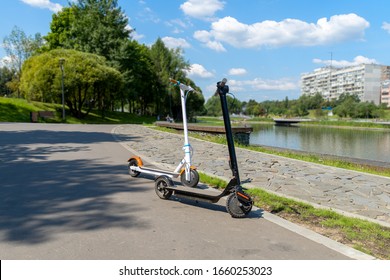 Two electric scooters, white and black, stand on the bandwagon on the street. Sunny summer day. Modern city transport. City park near the river. - Shutterstock ID 1660253023