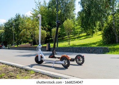Two electric scooters, white and black, stand on the bandwagon on the street. Sunny summer day. Modern city transport. City park with trees. - Shutterstock ID 1668254047
