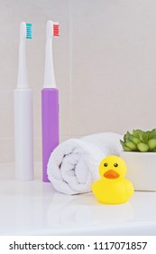 Two Electric Toothbrushes On Sink In The Bathroom With White Wrapped Towel And Yellow Rubber Ducky Duck