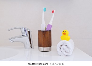 Two Electric Toothbrushes In Wooden Glass On Sink In The Bathroom With White Wrapped Towel And Yellow Rubber Ducky Duck. Personal Hygiene Concept. 