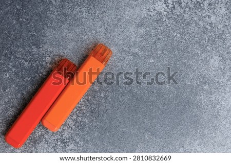 Two electronic cigarettes in red and orange colors are resting on a gray stone surface. They are positioned at an angle and the daylight highlights their design.