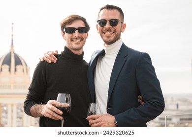 Two elegant men posing in front of natural white background looking at camera and smiling. They wear sunglasses and hold a glass of wine, on a rooftop terrace overlooking the city.