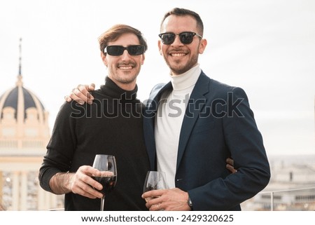Two elegant men posing in front of natural white background looking at camera and smiling. They wear sunglasses and hold a glass of wine, on a rooftop terrace overlooking the city.