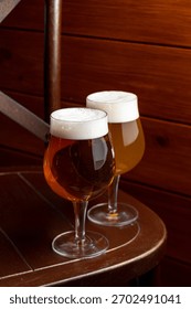 Two elegantly shaped glasses filled with craft beer rest on a wooden table in a cozy setting. The warm lighting enhances the inviting atmosphere, perfect for socializing and exploration