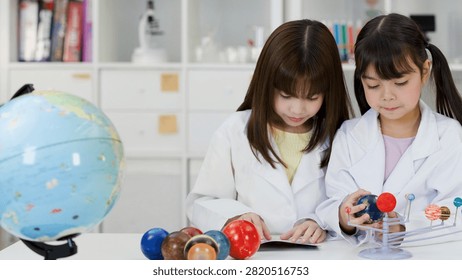 Two elementary school students in white lab coats examine a planetary model in a classroom.