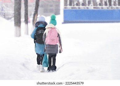 Two elementary schoolgirls go to school on a snow-covered path among snowdrifts. Snow is falling. Copy space.                               