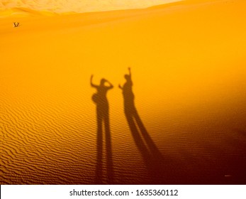 Two Elongated Black Shadows Of People Waving Against The Rippled Golden Sand Of  Arabian Desert Dunes On A Bright Sunny Day.  Relaxation And Vacation Concept.  Copy Space.