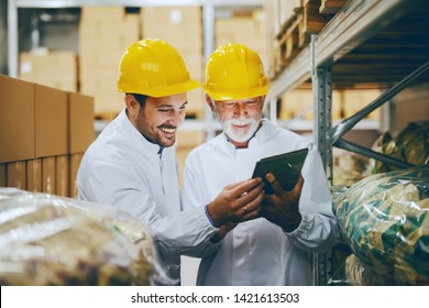 Two Employees Dressed In White Uniforms And With Yellow Helmets On Heads Checking Goods In Warehouse. Older Employee Holding Tablet And Talking To Younger One.