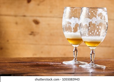 Two emptied glasses of beer on a counter with remnants of the lager and froth in the glass and copy space on a wooden background