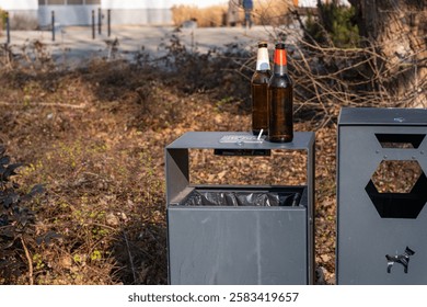 Two empty beer bottles with a cigarette butt rest on top of a modern outdoor trash bin in a public park taken in natural daylight with warmer tones.