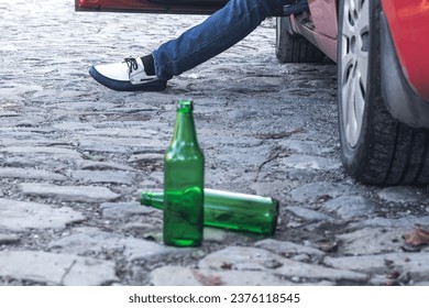 Two empty beer bottles on the floor next to the car.the driver of the car sits with the car door open.only his leg is visible. don't drink and drive concept.