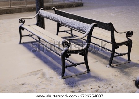Two empty benches sit covered in a fresh layer of snow during the winter night.