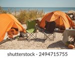 Two empty camping chairs set up by orange tents at a beachside campsite, awaiting campers. A peaceful and sunny coastal setting.
