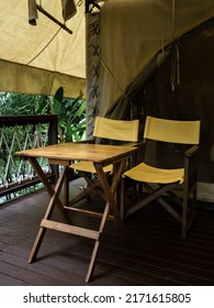 Two empty camping wooden chairs with yellow fabric seats and backrest and wood table on the terrace in front of the room of large tent. Folding outdoor chairs on white glamping tent, vertical style.