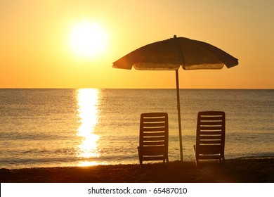 Two empty chairs stand on  beach under  opened umbrella with  view on marine sunset