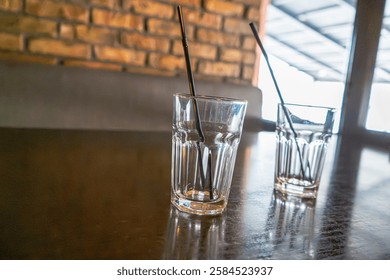Two empty glasses with black straws sit on a reflective wooden table in a cozy café or restaurant setting. The background features a warm brick wall and large windows allowing natural light in