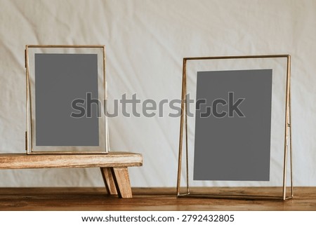 Two empty gold-framed photo frames on a wooden surface against a neutral background, one standing on a small wooden platform.