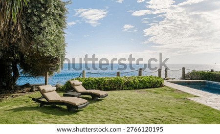 Two empty lounge chairs on a manicured lawn overlooking a calm blue ocean, next to a private pool and tropical greenery