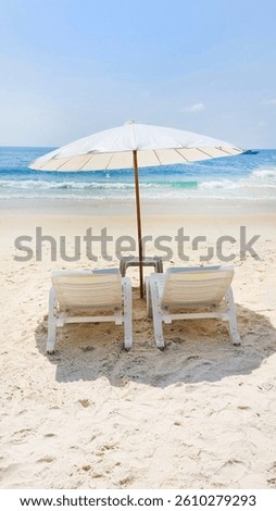 Two empty white beach chairs standing under a white parasol on a tropical beach with turquoise water and white sand