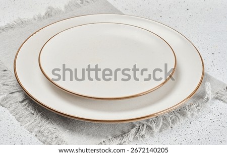 Two empty white ceramic plates with brown rims stacked on a gray linen napkin with fringe