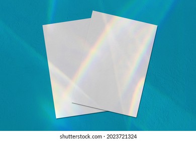 Two empty white vertical rectangle a4 poster, business card mockups lying diagonally on top of each other with overlay of rainbow light refraction caustic effect on mint blue concrete background