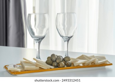 Two empty wine glasses and a serving tray with assorted cheeses and green olives on a white table. The background features soft natural lighting and a neutral curtain, creating a refined aesthetic - Powered by Shutterstock